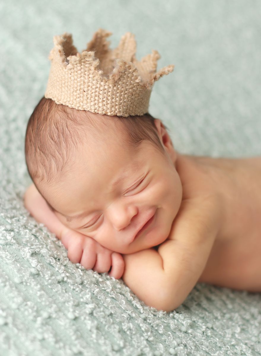 Profesionální fotografie spícího novorozeňátka s korunkou na hlavě, rukama pod bradou a šibalským úsměvem
