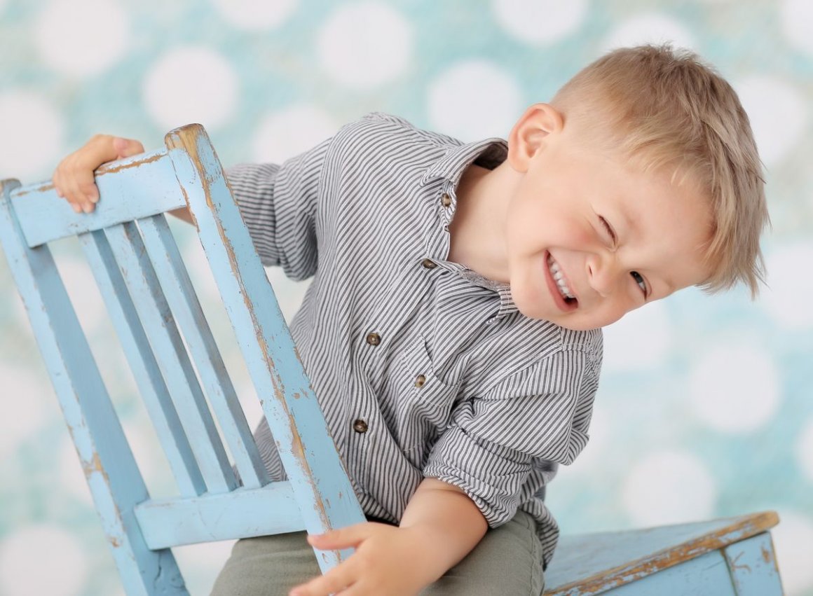 Profesionální fotka pětiletého blonďatého chlapce dovádějícího na modré otlučené dřevěné židli ve fotoateliéru před barevným fotopozadím. Chlapec vesele mrká a na židli se zřejmě kývá ze strany na stranu.