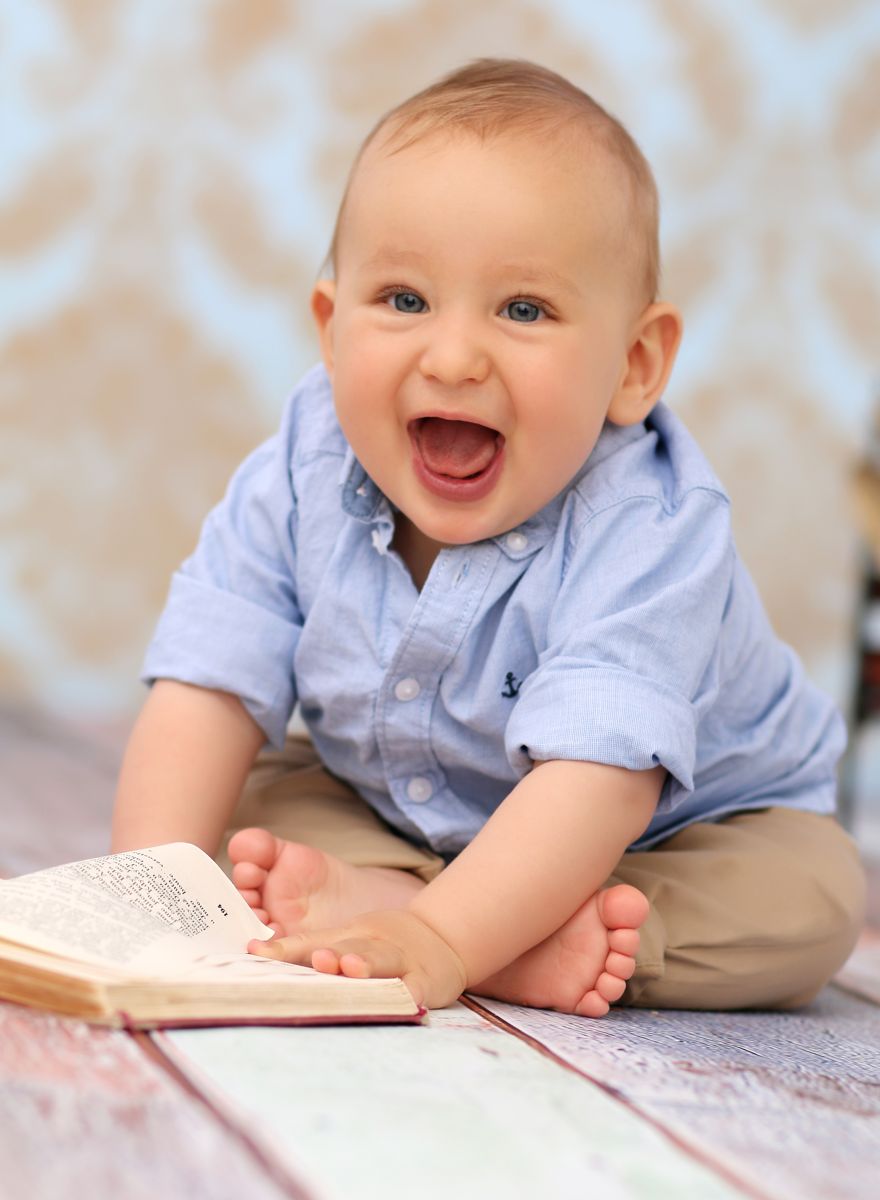 Půlroční miminko, chlapec v modré košili na profesionálním focení se šťastně směje a rukou na podlaze listuje starou knížkou. | © Fotoprome.cz