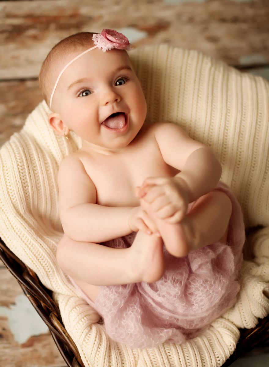 Půlroční miminko ve fotoateliéru (holčička) s květinovou čelenkou leží na zádech, drží ručičkami za nožičky a na plnou pusu se směje. | © Fotoprome.cz