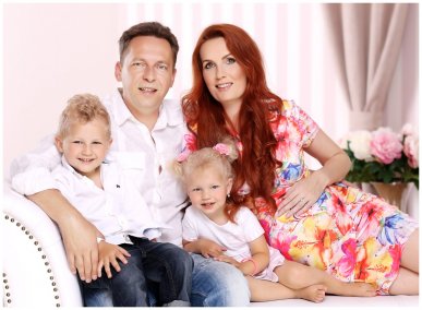 Těhotná maminka s tatínkem a dvěma dětmi sedí v ateliéru na bílém chaise longue, maminka má květované šaty a dlouhé zrzavé vlasy, ostatní mají bílé košile a jeansy.