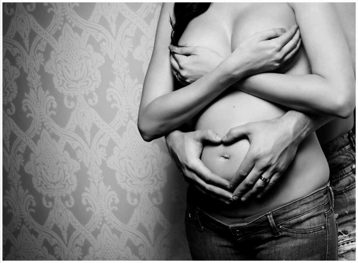 Detailní černobílá fotografie těhotenského bříška, na kterém ruce maminky a tatínka tvoří symbol srdce. Intimní a vkusný portrét polonahých rodičů na pozadí s výraznými tapetami.